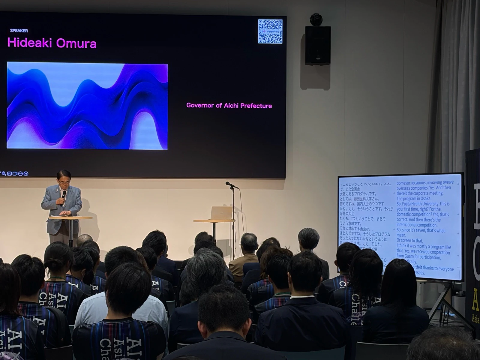 Aichi Prefecture Governor Hideaki Omura speaking at AI×MedTech, with a VoicePing QR code on the main screen and Japanese–English live captions on a side display.