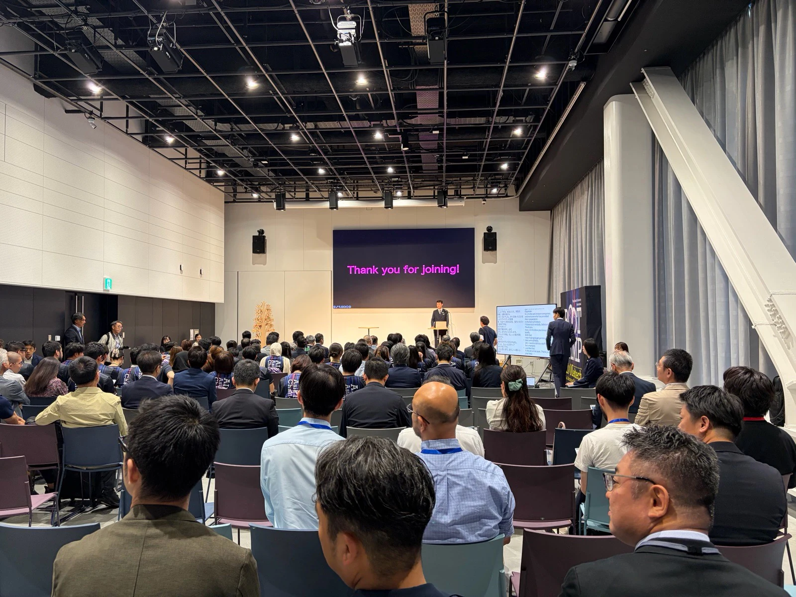 Audience facing the AI×MedTech stage and adjacent bilingual caption screen at STATION Ai.