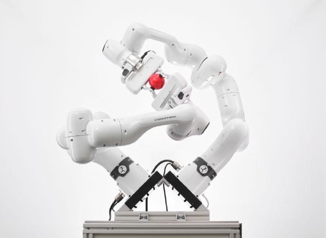 Two complete Franka Research 3 arms mounted on an angled stand and holding a red object together; each arm has seven degrees of freedom.
